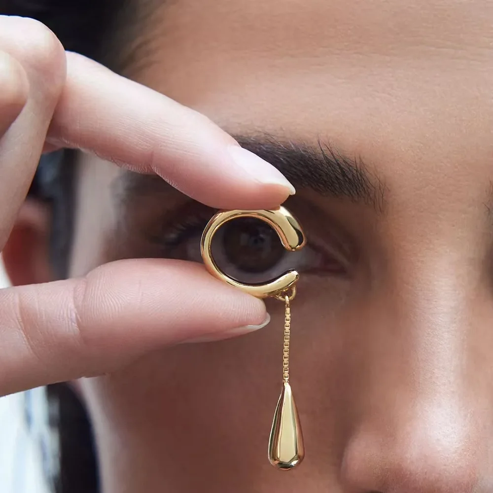 Clip d'oreille doré en C tenu entre les doigts, goutte pendante